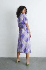 Sampa Dress in a purple and white checkered print, knee-length with waist tie, paired with green sandals against white wall.