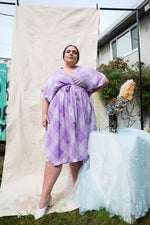 Wrap-front Sampa Dress in purple Eostre print, tied at waist, set against fabric backdrop with a rustic table arrangement.