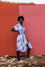 Chic Sampa Dress in purple Eostre print, with a waist belt and white shoes, against a red wall with natural elements.