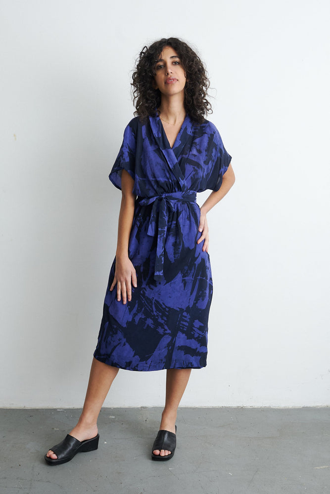 Blue Sampa Dress with abstract pattern, tie waist, short sleeves, black sandals, against light wall, industrial setting.,