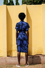 Back view of Sampa Dress in Rorschach print, blue with white pattern, short curly hair, standing against worn yellow wall.