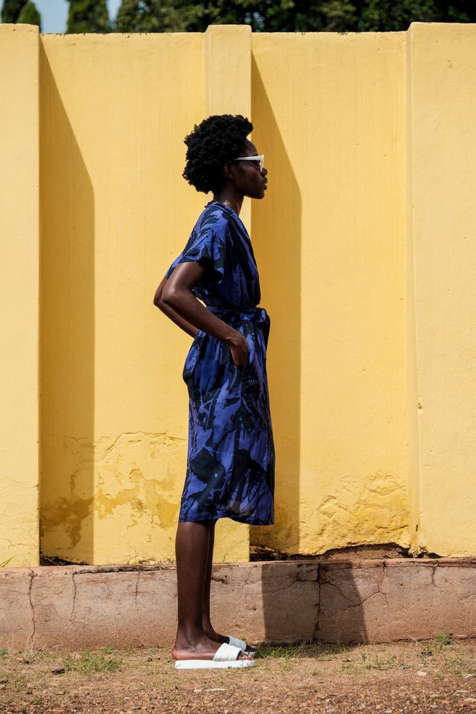 Casual stance in Sampa Dress with Rorschach print, waist-tied, paired with white slip-on shoes, against a yellow wall.