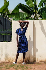 Person in Sampa Dress with blue and white Rorschach print, short sleeves, waist tie, against cream wall with green plant.