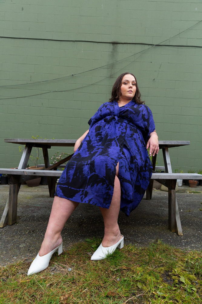 Chic blue batiked Sampa Dress with abstract pattern, worn on a wooden bench, paired with white heels, in an outdoor setting.