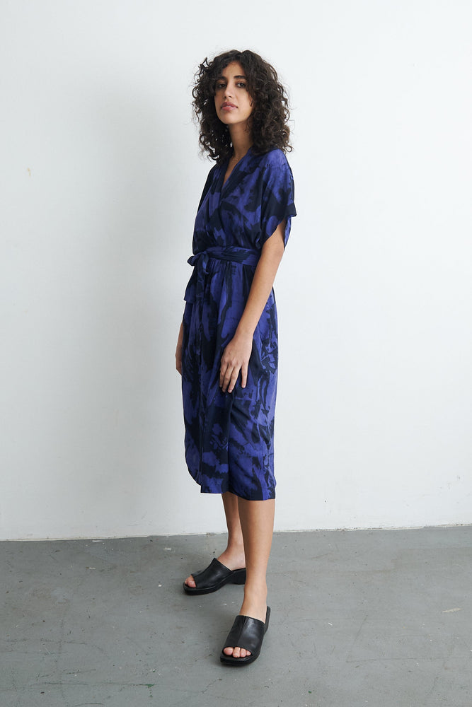 Blue Sampa Dress with abstract print, shoulder-length curly hair, black shoes, light wall backdrop.