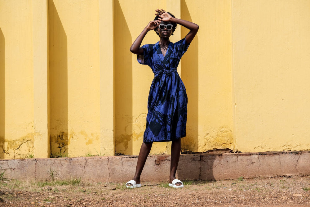 Blue Sampa Dress with tie waist and short sleeves, white pattern, standing on red dirt ground, yellow wall backdrop.