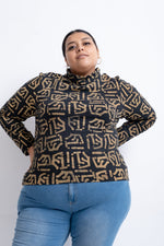 Person in a high-neck, long-sleeved top with a geometric pattern, paired with blue jeans against a white backdrop.