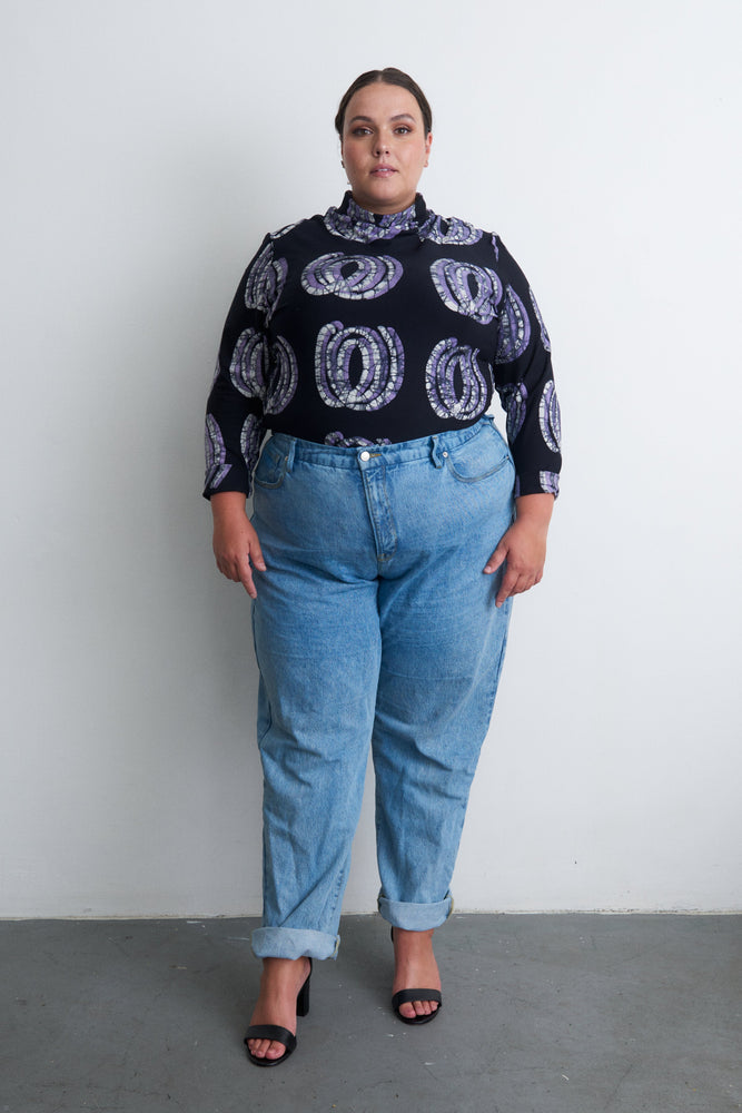 Person in the Stricta Turtleneck in Good Signal and jeans, against a white wall, focus on the unique, hand-batiked top.