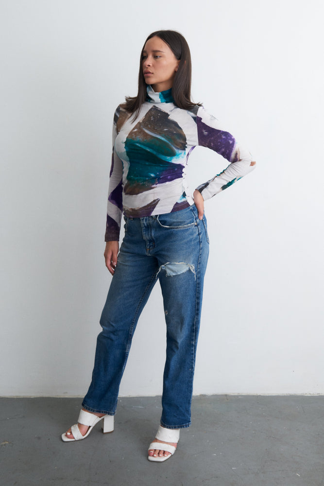 Confident pose in Osei-Duro abstract patterned top and distressed jeans, with white heeled sandals, against a light wall.