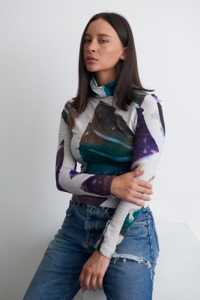 Abstract blue, purple and green patterned top with distressed jeans, crossed arms pose, against a white backdrop.