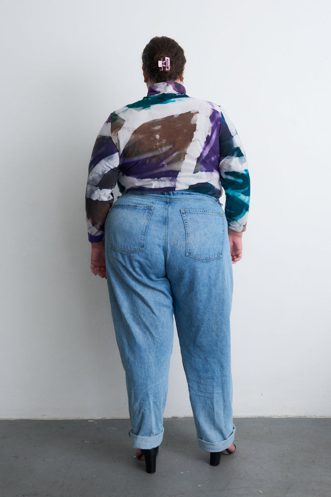 Rear view of person in multicolored patterned shirt and blue jeans, with a hair clip, standing against a white wall.