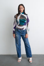 Abstract multicolored Stricta Turtleneck paired with blue jeans and white sandals, against a light wall.