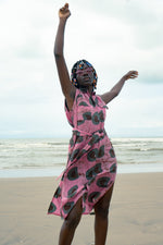 A woman wearing a pink dress with a green and black print, stands on a beach with one arm raised in celebration or triumph.