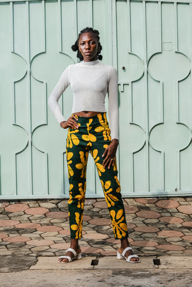 Model in front of patterned blue doors, wearing Vitta Trousers with green and yellow design, paired with white sandals.