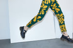 Legs in Vitta Trousers step through white panel, showcasing bold yellow leaf print and black loafers.