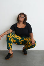 Model sits against white wall in Vitta Trousers, showcasing yellow floral design and relaxed fit with visible tattoo.