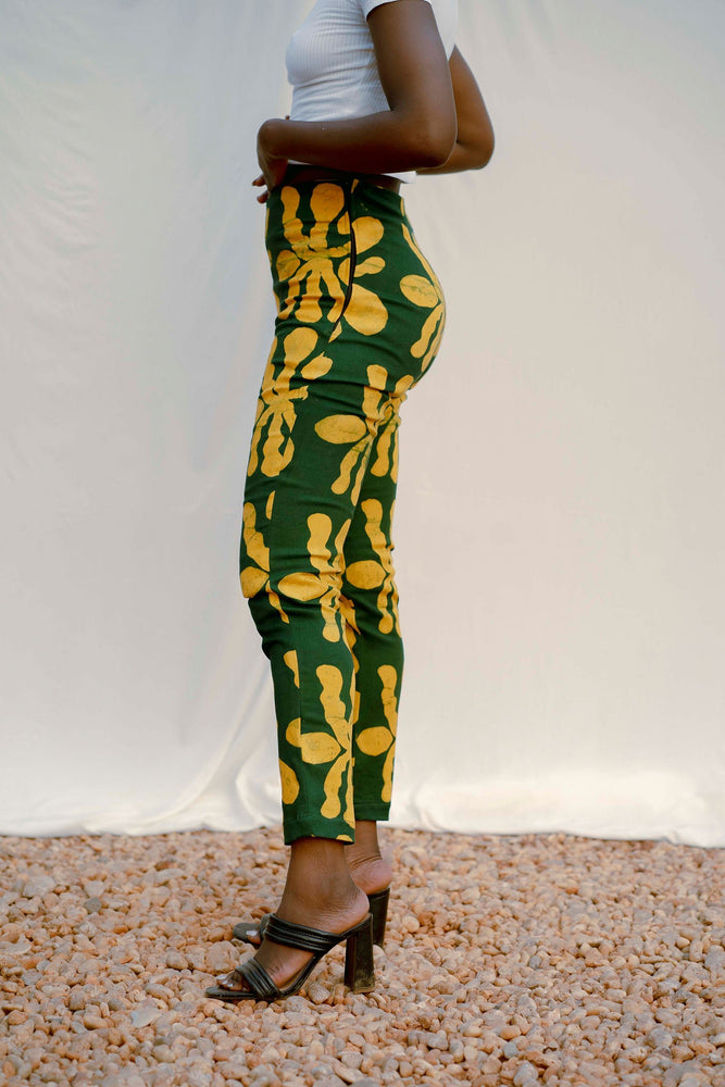 Lower half of model in Vitta Trousers with green leaf pattern, paired with black sandals, standing on gravel.