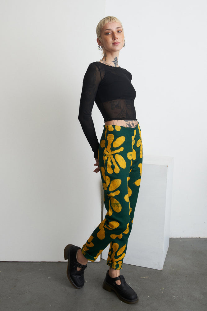 Model stands against white backdrop, black sheer top, Vitta Trousers with green and yellow floral print, black shoes.