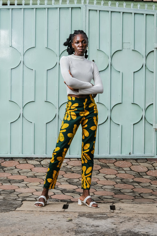 Model in Vitta Trousers by light green gate, white top and sandals, showcasing yellow-green floral design.