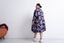 Person in floral Para Dress facing away, bun hairstyle, white shoes, beside orange ottoman on light floor.