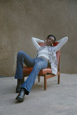 Relaxed individual in zebra like Long Division print Index Vest and blue jeans, sitting on a brown chair in an outdoor setting.