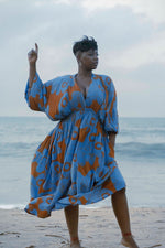 Person in All Ideas print Bating dress stands on beach, hand raised, against calm ocean and overcast sky.