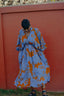 Person in a vibrant All Ideas print Bating Dress against a red wall, with dark hair and strappy sandals.