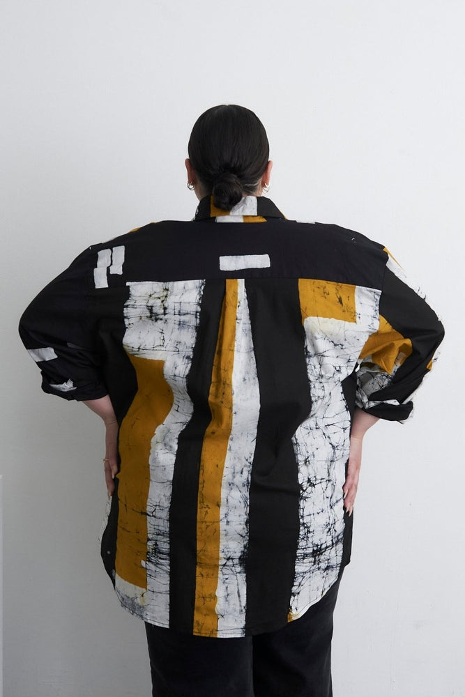 Back view of the Bula Shirt with black, white, yellow abstract print, layered over black jeans, against a white background.