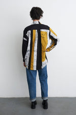 Artistic Bula Shirt with abstract black/white/yellow design, paired with rolled-up jeans and black shoes, against a white wall.