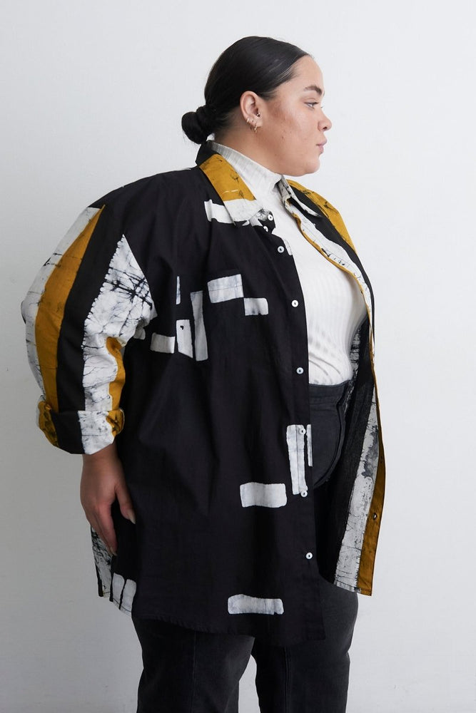 Person in artistic Bula Shirt with black, white, yellow abstract print, layered over white and yellow top, against a white background.