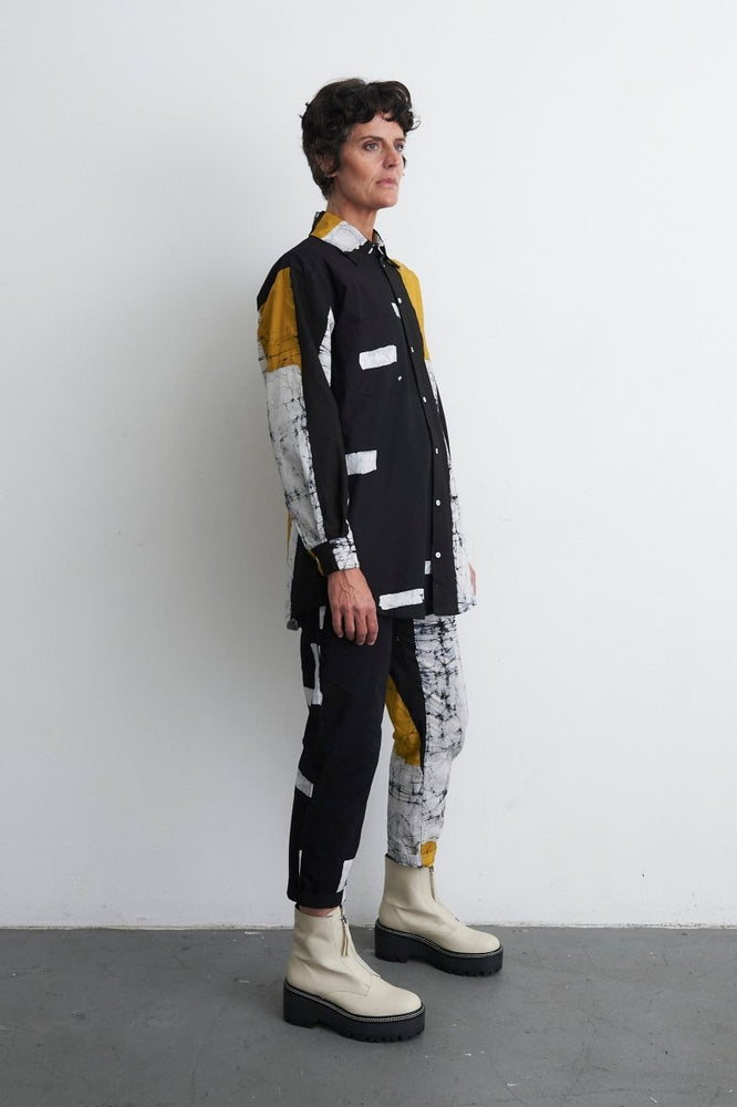 Avant-garde Bula Shirt with abstract black/white/yellow design, paired with Kpong trousers and chunky beige boots, against a white wall.