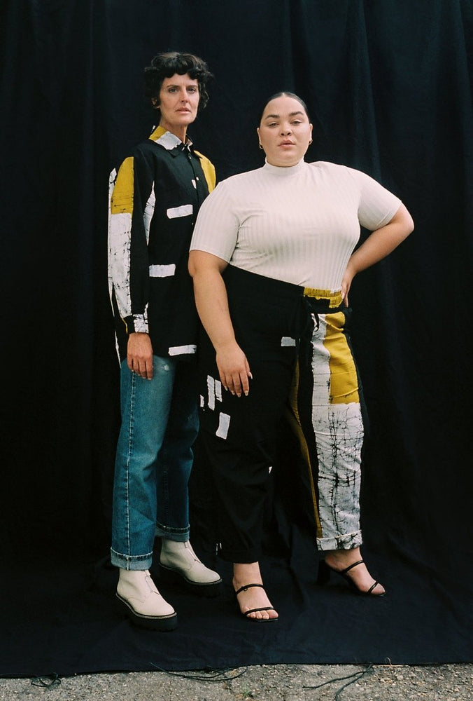 Two individuals in abstract Bula Shirts against a black backdrop, one with white boots and the other with black heels, exuding style.