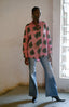 Woman standing indoors in pink Bula Shirt with avocado-like print, flared denim trousers, black pointed-toe heels.