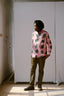 Man standing in pink Bula Shirt with green and black abstract patterns, olive green trousers, black shoes, indoors.