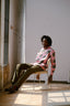 Man sitting on a chair next to a window, wearing the Bula Shirt in Tunnel of Love, a batik print shirt, trousers, and shoes.