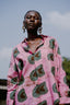 Woman wearing a pink Bula Shirt with bold green and brown patterns, front pocket, white buttons, standing outdoors.