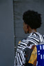 Person in a colorful Bula Shirt with stripes and abstract shapes, standing against a textured dark grey wall, exuding urban chic.