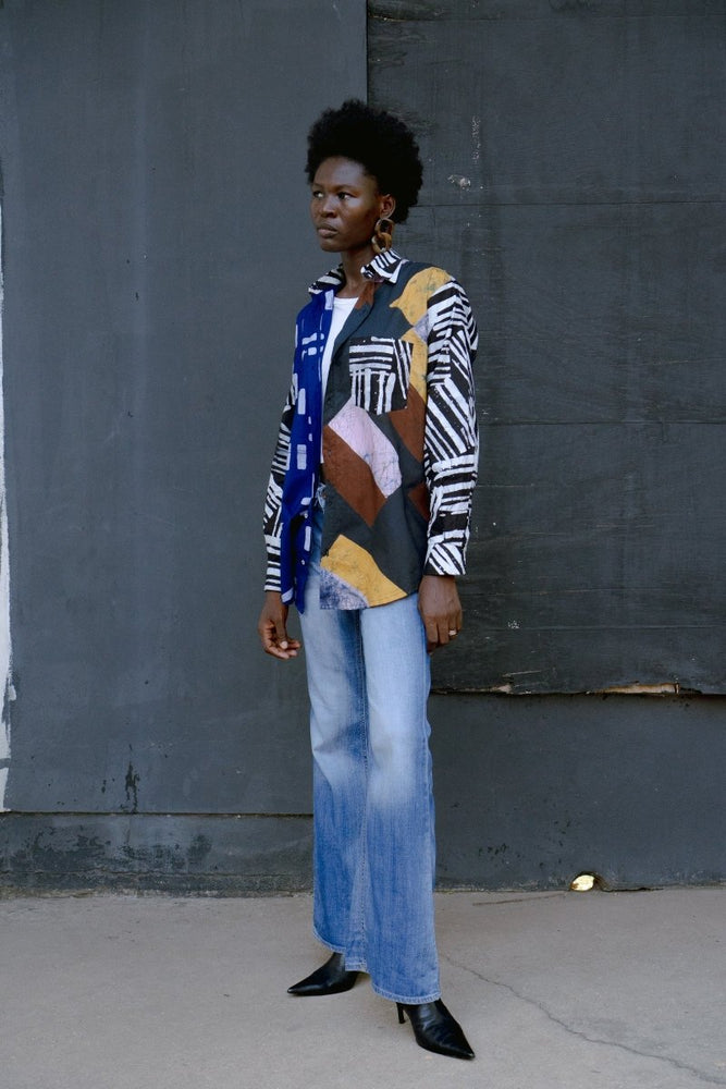 Fashion-forward Bula Shirt with blue, white, and brown prints, paired with faded jeans and black shoes, against a grey wall.