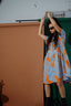 Helia Dress in All Ideas print, abstract orange flowers on sky blue, with sunglasses on, against a textured wall backdrop.