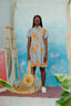 Person in Helia Dress with orange-blue print, holding a straw hat, standing by a blue-white canvas and wooden easel.