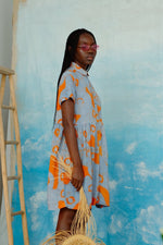 Knee-length Helia Dress in patterned orange and blue, model is in front of a blue textured wall with a wooden ladder.