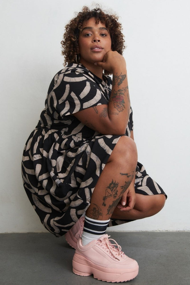 Person in Cantaloop print Helia Dress sits against white wall, tattoos and pink platform sneakers on show.