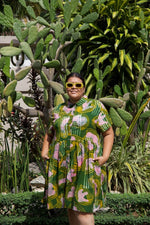 Green Helia Dress with pink-white print, model wears sunglasses, in a garden with cacti and lush plants.