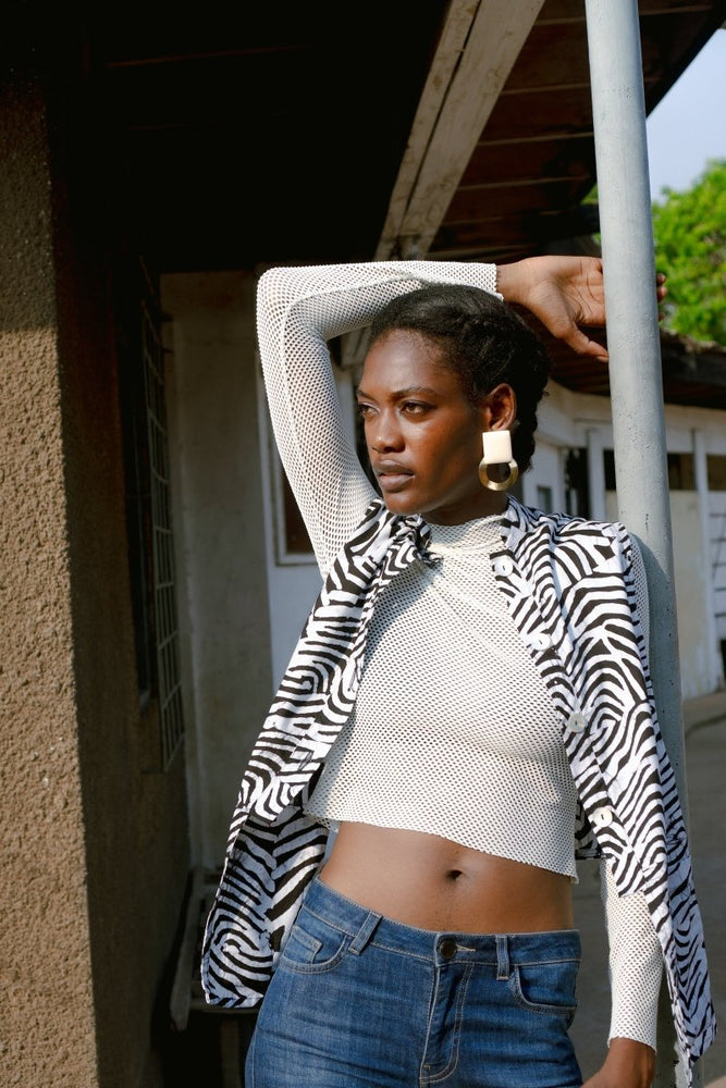 Person in Long Division print top and high-waisted jeans, standing outdoors under a structure, hand on head.