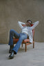 Relaxed individual in zebra like Long Division print Index Vest and blue jeans, sitting on a brown chair in an outdoor setting.