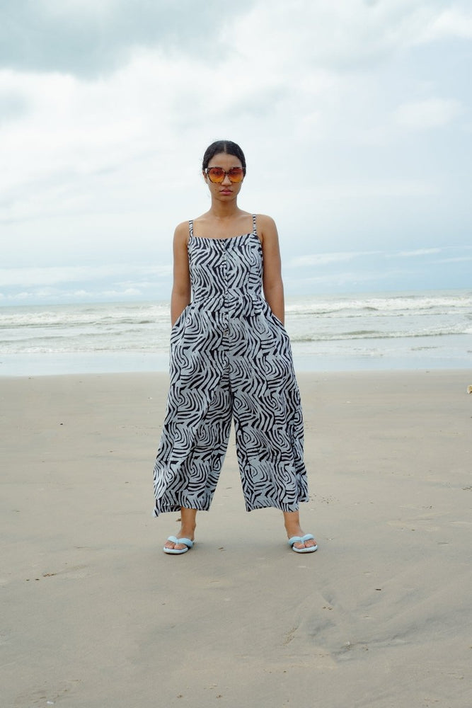 Sleeveless Mafalda jumpsuit with black & white wavy print, styled with silver sandals on a sandy beach backdrop.