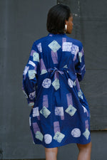 Back view of woman in blue Press Dress in Location with geometric pattern of rectangles and circles.