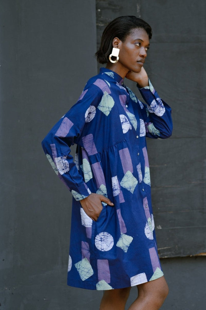 Side view of woman in blue Press Dress with purple and white geometric patterns, hand on hip, wearing gold earrings.