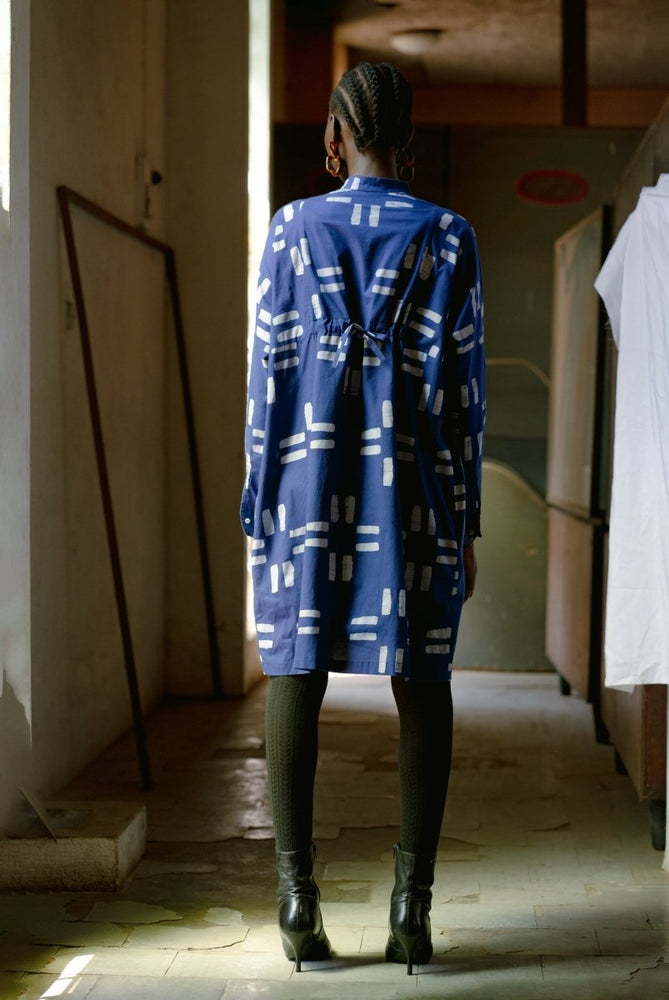 Back view of model in Press Dress with white patterns and black boots, standing in a corridor with natural light.