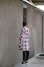 Back view of a model in a pink Press Dress with green batik print, standing outside a textured building with a barred window.